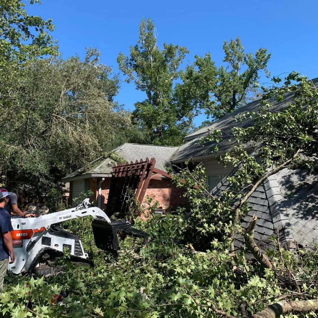 A worker from Northshore Tree Storm Prep & Risk Mitigation operates a Bobcat skid-steer loader to remove fallen branches from a house’s roof, covered with debris after a storm. Large trees and blue sky are visible in the background.