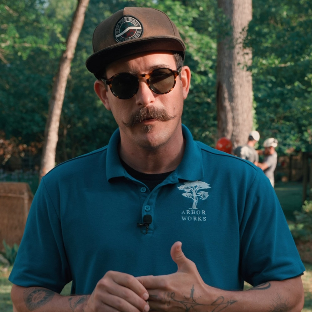 A man wearing sunglasses, a baseball cap, and a blue "Arbor Works" polo shirt stands outdoors in a wooded area, looking at the camera. The image is perfect for an Elementor Single Post background with blurred people and trees behind him.