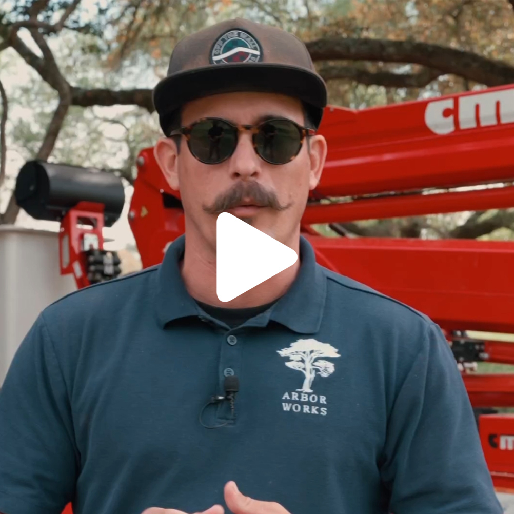 A man with a mustache, wearing sunglasses, a dark cap, and a blue Arbor Works shirt stands outdoors in front of red Northshore Tree Removal machinery. Trees are visible in the background. A play button icon is overlaid on the image.