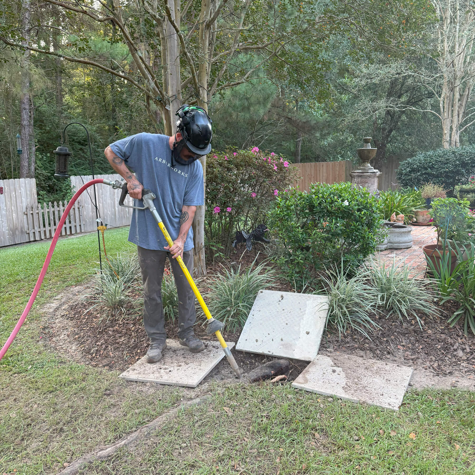 A person wearing a helmet and face shield uses equipment to work on an underground utility access point in a landscaped backyard, possibly for Root Pruning Northshore. Concrete slabs are removed, with various tools and a hose also present.