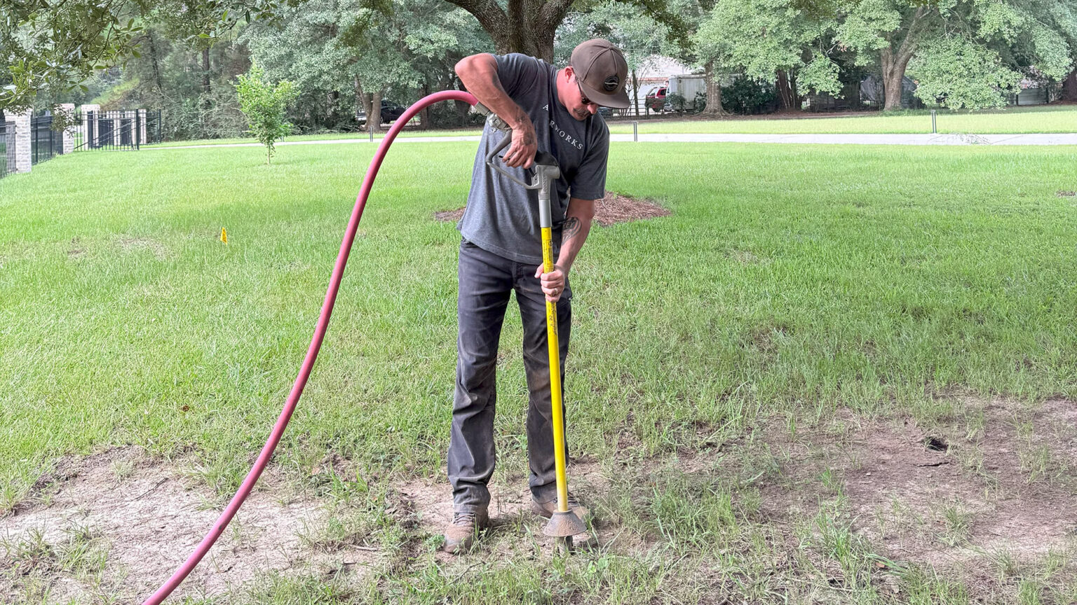 A person stands on grass using a yellow digging tool with one hand and holding a red hose with the other, performing vertical mulching Northshore, surrounded by trees and a fenced yard in the background.