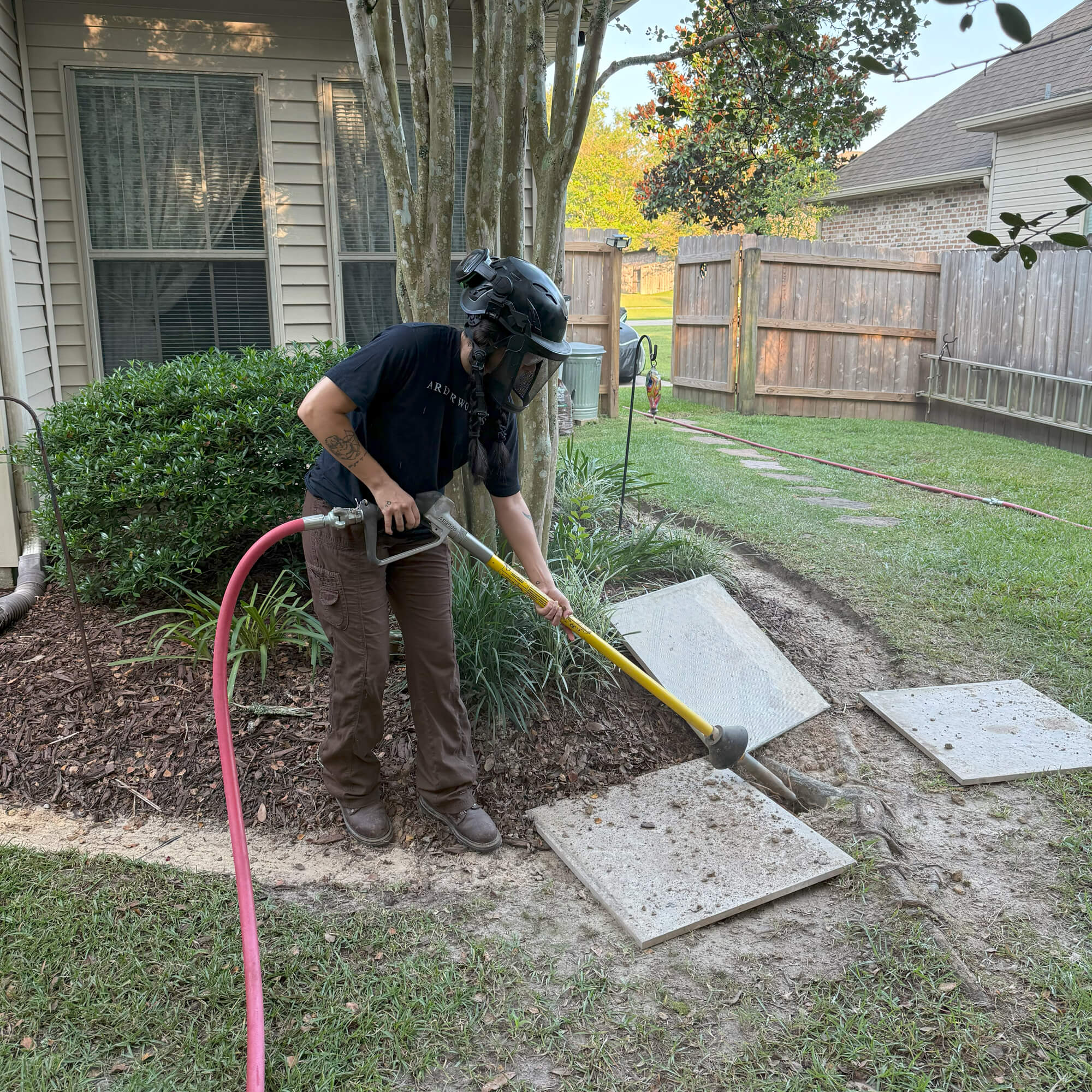 A person wearing protective gear uses a tool connected to a pink hose to clear dirt around concrete pavers near a house, performing Root Pruning Northshore-style, with a shrub and some trees visible in the yard.