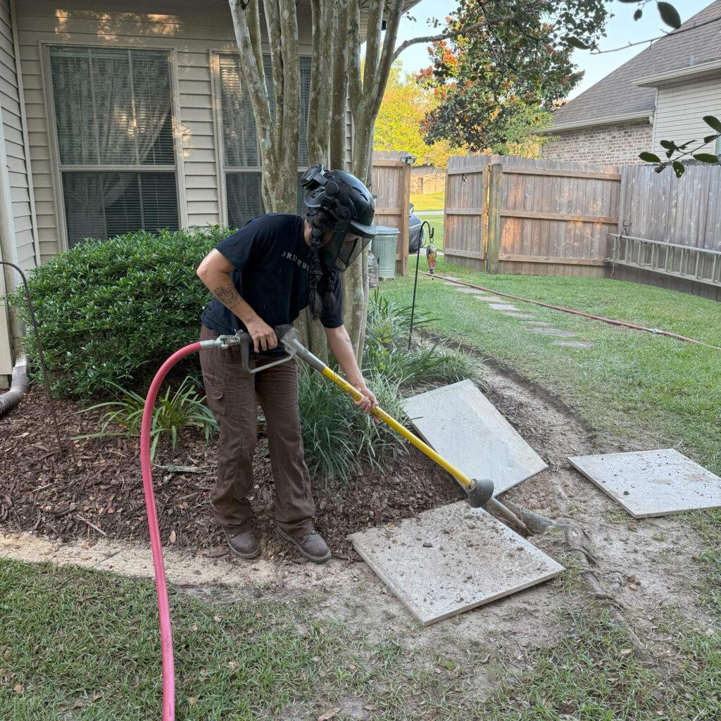 A person wearing protective gear uses a tool to remove dirt from concrete stepping stones next to a house, possibly as part of root pruning Northshore services, with a garden bed, bushes, and a wooden fence in the background.