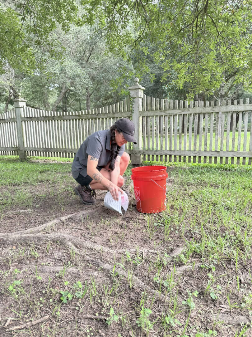 A person wearing a cap and shorts is crouching on the grass near a wooden fence, holding a white container beside an orange bucket—possibly preparing for Vertical Mulching Northshore. Trees and a grassy area are visible in the background.