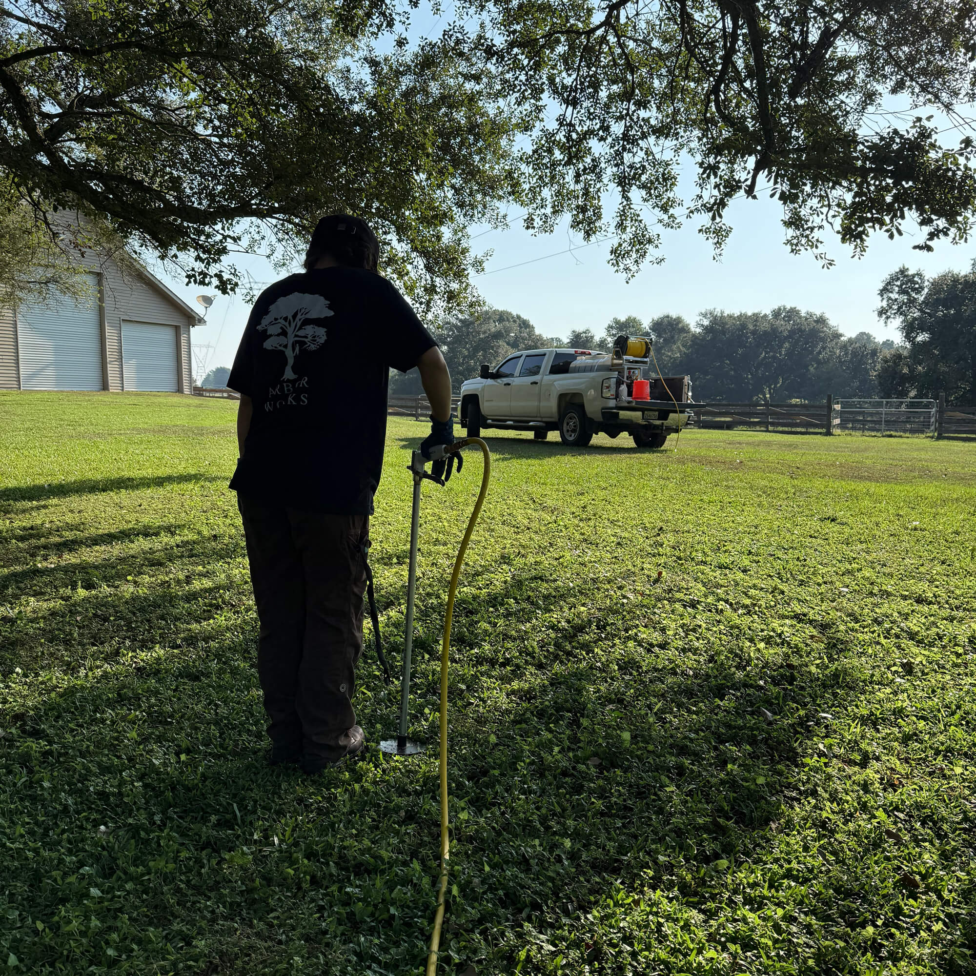 A person uses a soil probe for Deep Root Fertilization on a grassy lawn near a large tree, facing away from the camera. A pickup truck with equipment is parked in the background near a building with a white garage door.