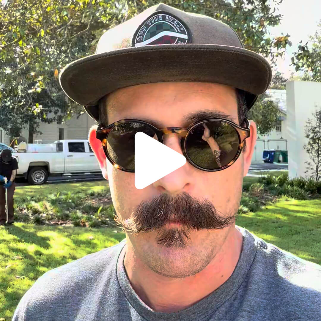 A man with a large curled mustache, sunglasses, baseball cap, and gray shirt stands outside on a sunny day, possibly discussing Deep Root Fertilization. A white pickup truck and greenery are in the background with a play button overlaying the center.