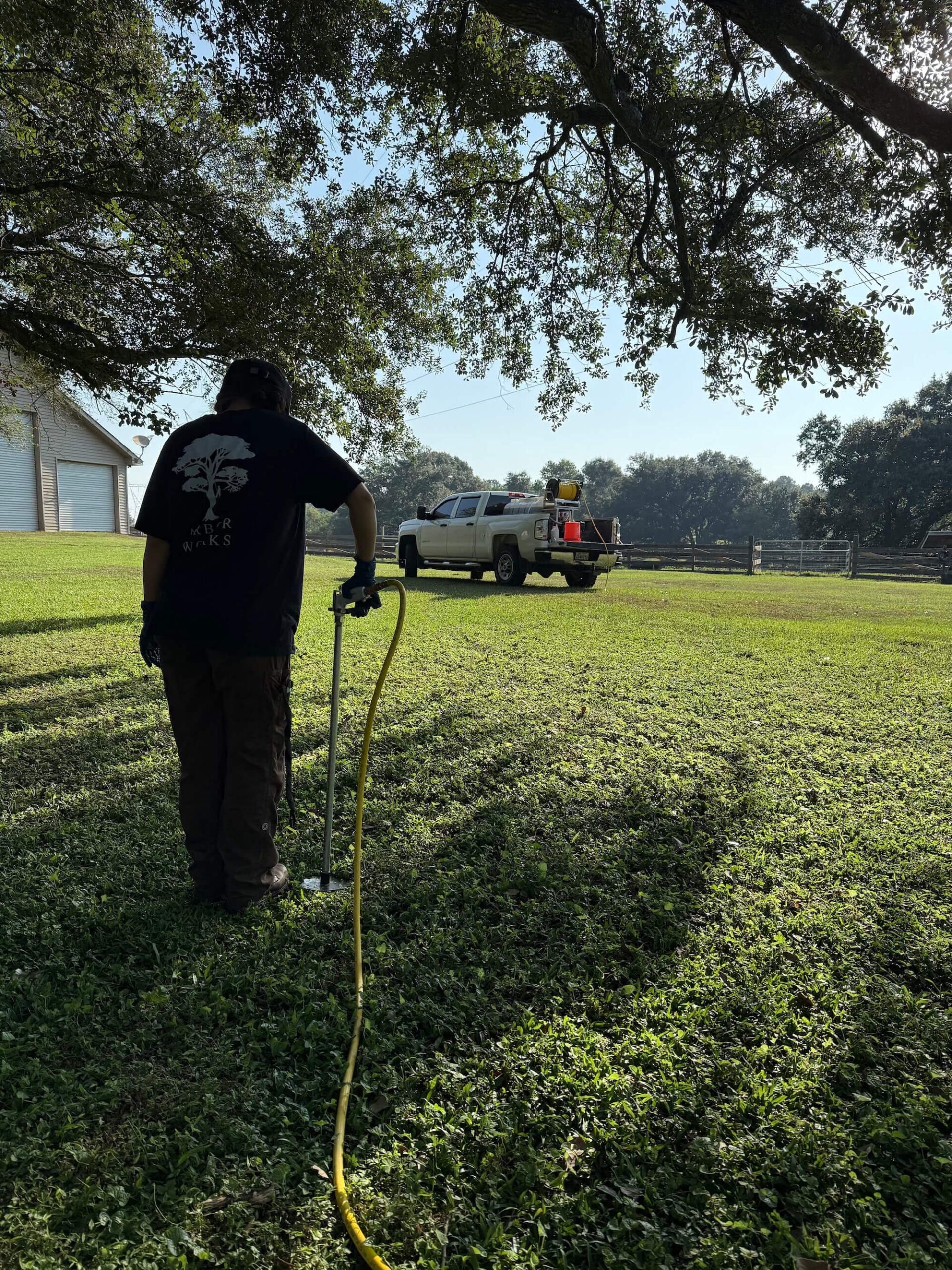 A person stands on a grassy lawn using a tool connected to a yellow hose, performing tree fertilization Northshore. In the background, a white pickup truck is parked near a fence and building, with trees providing shade.
