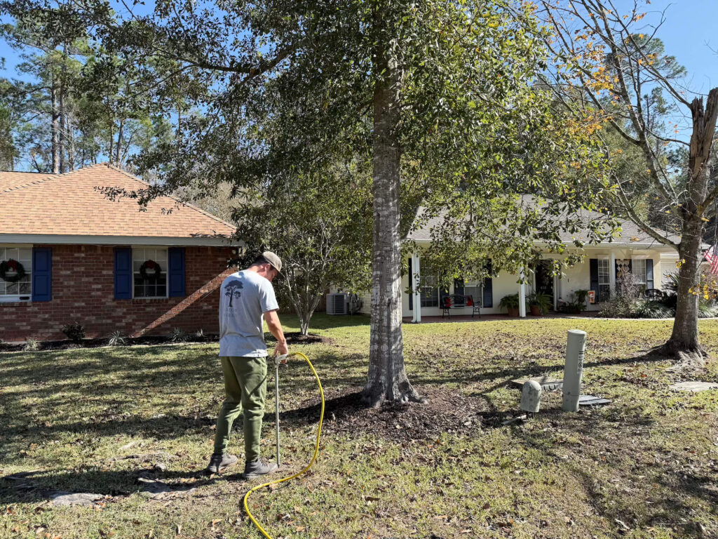 A person in a gray shirt and green pants uses a yellow garden hose near the base of a tree, demonstrating Tree Fertilization Northshore techniques, with two houses and trees visible in the sunny front yard background.