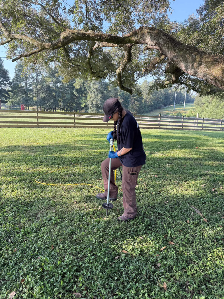 A person wearing gloves, a hat, and outdoor clothing uses a soil probe for tree fertilization in a grassy Northshore yard with a large tree and wooden fence in the background.
